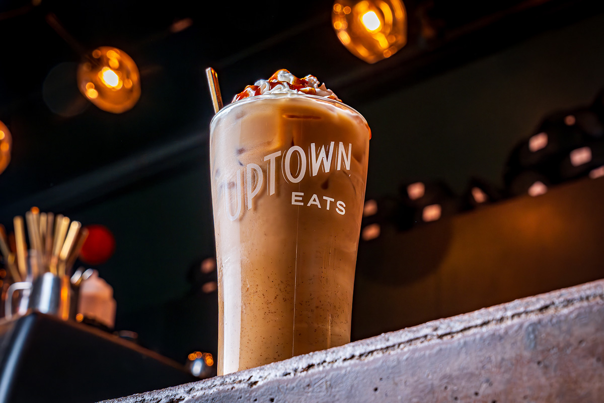 A tall iced coffee cooler in an Uptown Eats cup on a wooden counter