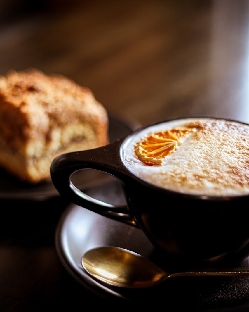 A cappuccino dusted with cinnamon beside a cinnamon scone on a dark table