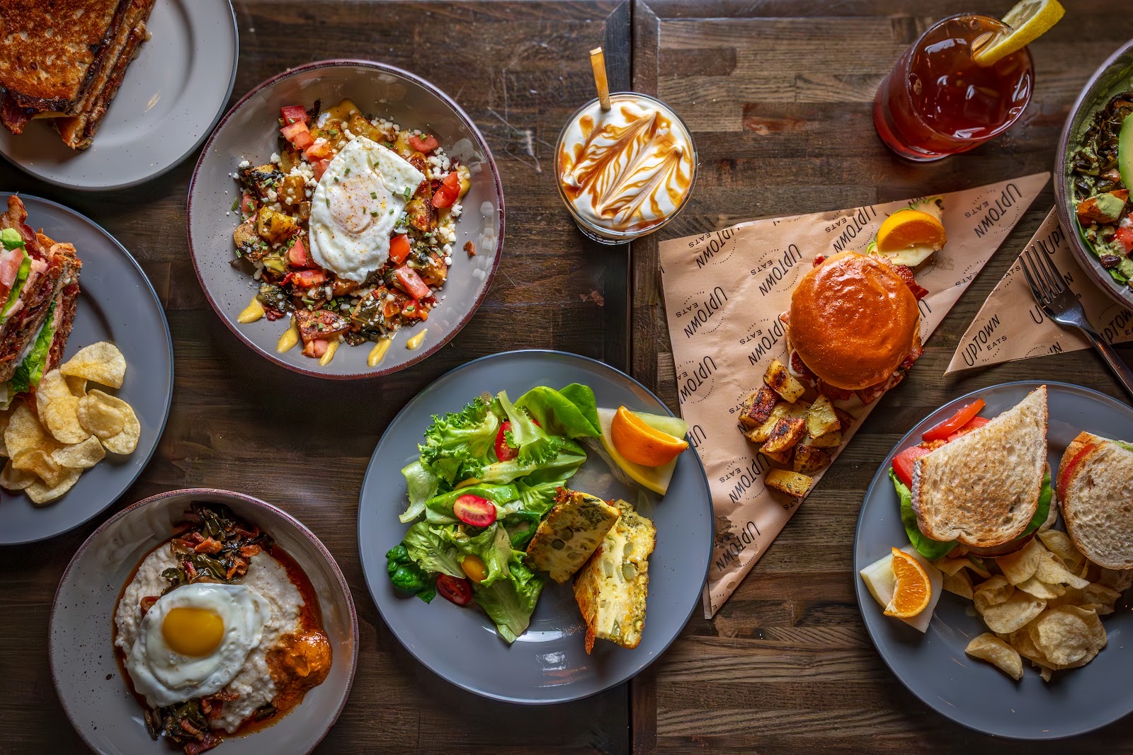 An overhead spread of brunch plates, a burger, a bowl and iced drinks on a wooden table